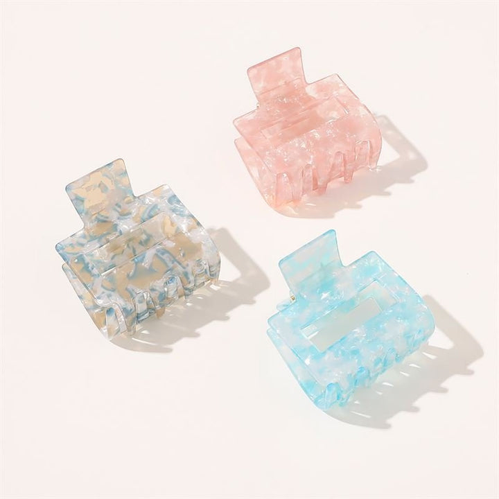 The SOHO Ema Hair Claw - Crystal Blue by SOHO features a translucent, marbled rectangular design with wide teeth, perfect for elegant hairstyles. Displayed alongside pastel pink and green, the clips rest on a light background.