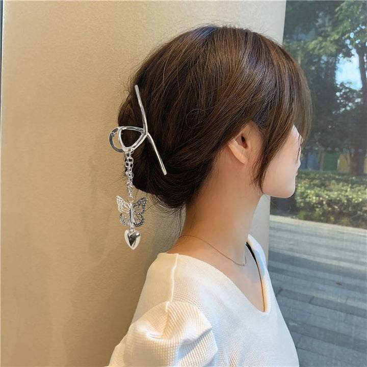 A woman with brown hair in a low bun, secured by the SOHO Emy Metal Hair Claw - Butterfly, wears a white top and stands by a window with trees outside.