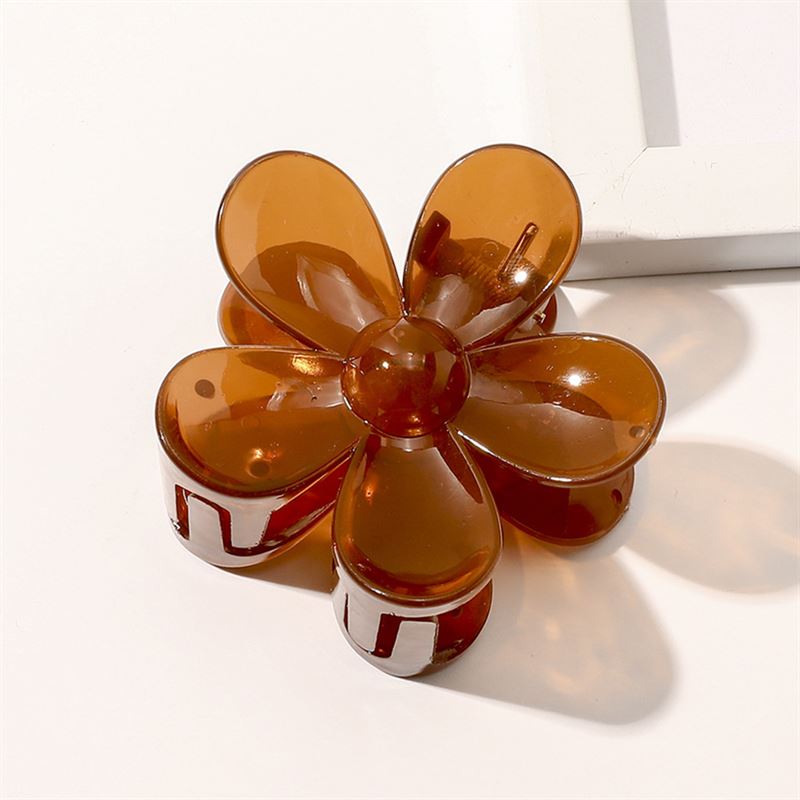 The SOHO Fiore Hair Claw - Russet, a brown translucent six-petal accessory from SOHO, is shown on a white surface near the corner of a white rectangular object.