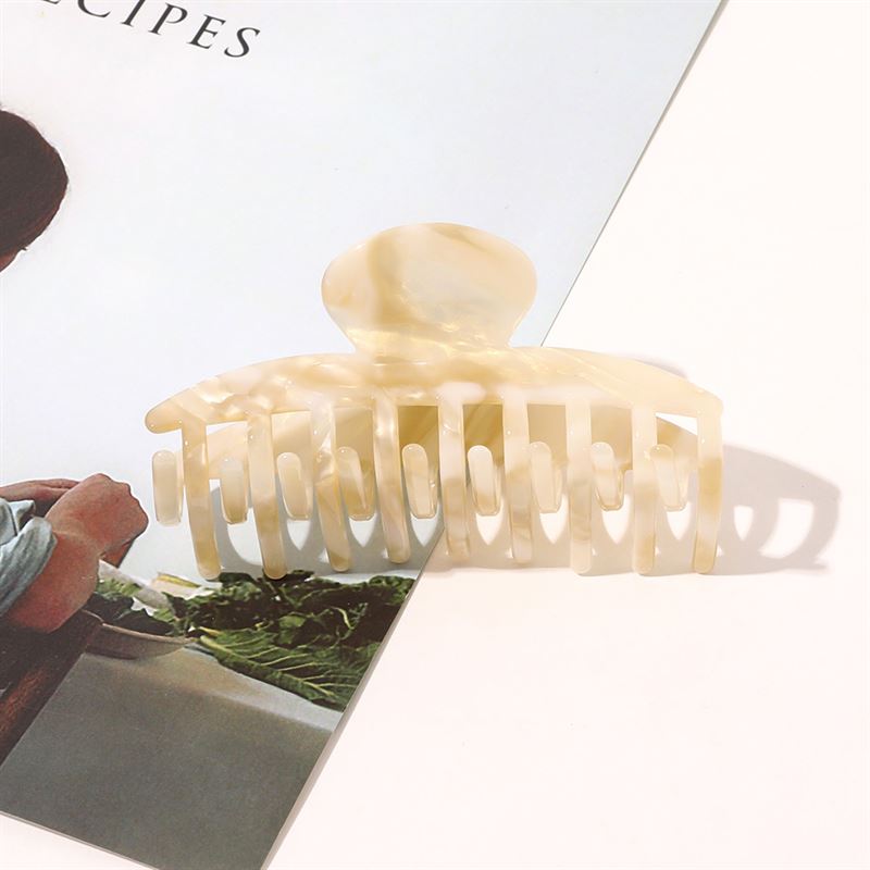 The SOHO Gaja Hair Claw in Beige Marble rests on a magazine, its elegant marbled design highlighted by soft shadows. The magazine's photo features greenery and a person, complementing the chic SOHO accessory.