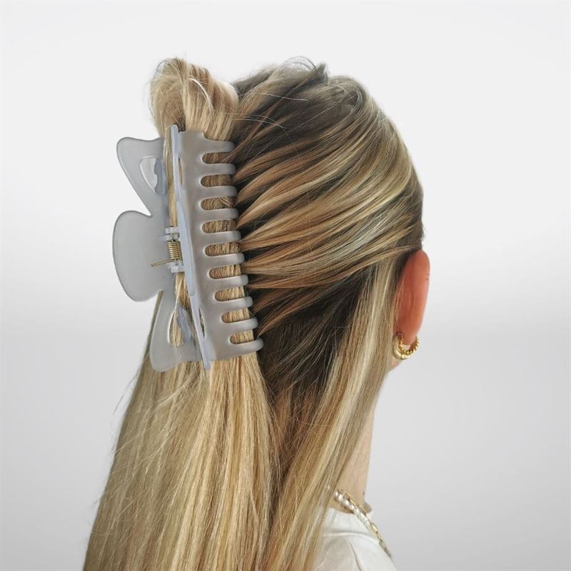 A woman with long blonde hair secures a half-up hairstyle using the SOHO Jelly Hair Claw in Cool Blue, showcasing its strong grip. Seen from behind against a plain background, she wears gold hoop earrings and a necklace.