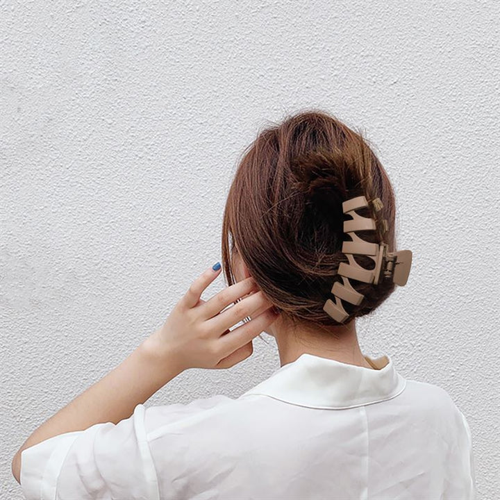 A person with brown hair in a bun, secured with the SOHO Just Hair Claw - Green by SOHO, wears a white collared shirt and stands facing a textured white wall, raising their hand near their face.