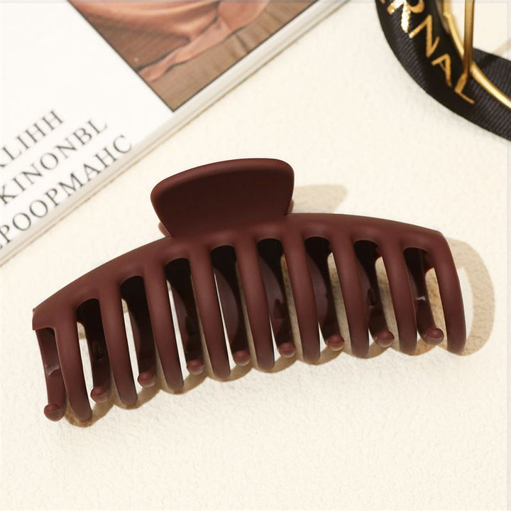 The SOHO Kana Hair Claw in Bark Brown, designed for all hair types, features a strong grip and matte finish. It rests on a light surface near a magazine and a gold-lettered ribbon.