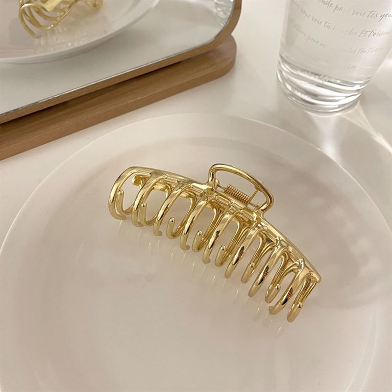 The SOHO Kana Metal Hair Claw in Gold by SOHO, designed for all hair types, features a strong grip and sits on a white plate with a mirror and clear glass of water in the background on a light surface.