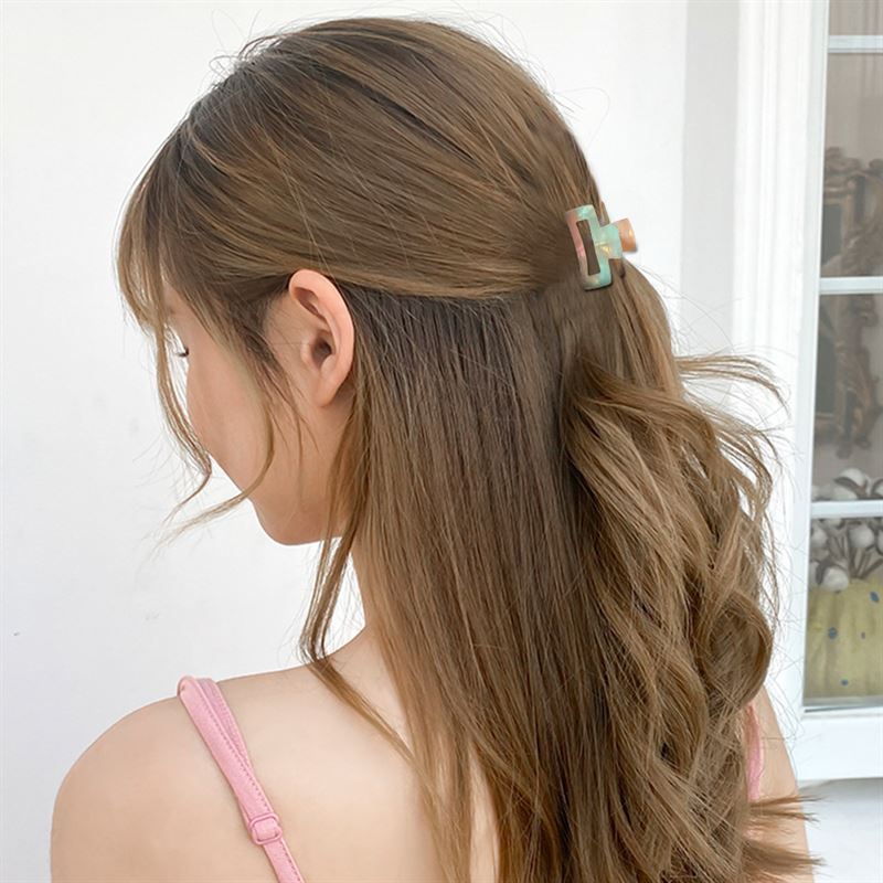 A woman with long, wavy brown hair wears a pink tank top and secures her hair with the SOHO Leia Mini Hair Claw in Tortoise. She is seen from behind against a white background with part of a window visible.