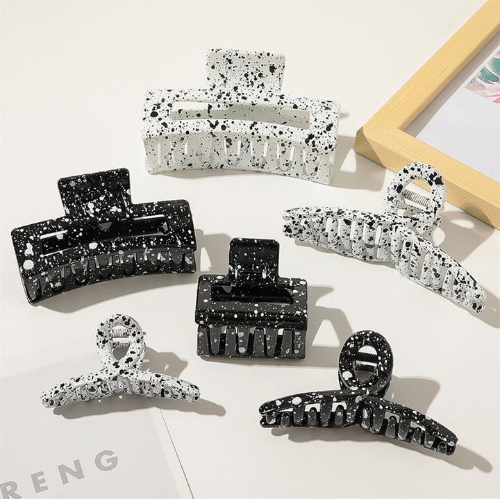 Six SOHO Luva Hair Claw clips in white, featuring sleek black and white splash patterns, are arranged on a white surface near a wood-framed picture. Each SOHO clip keeps your hair stylishly secured.