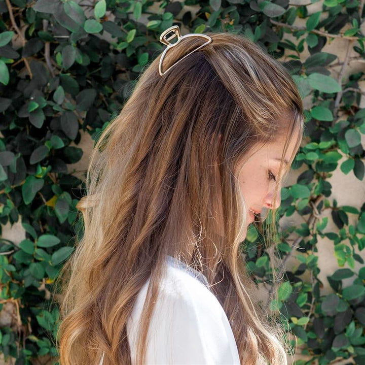 A woman with long, wavy light brown hair styled half-up with the SOHO Lyra Large Metal Hair Claw in Gold by SOHO stands in profile against leafy greenery, wearing a white top and smiling gently.