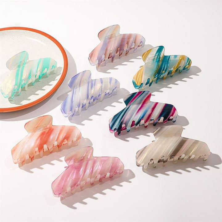 Seven SOHO Mesa Hair Claw - Grey Rainbow clips by SOHO are displayed on a white surface. A small round mirror in the corner reflects two of the hair claws, each featuring unique pastel or vibrant grey rainbow color patterns.