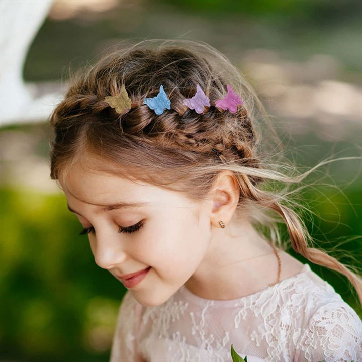 A young girl with braided hair adorned with SOHO Mini Butterfly Hair Claws (50 pcs, multicolour) smiles softly outdoors, wearing a light pink lace dress against a blurred green background.