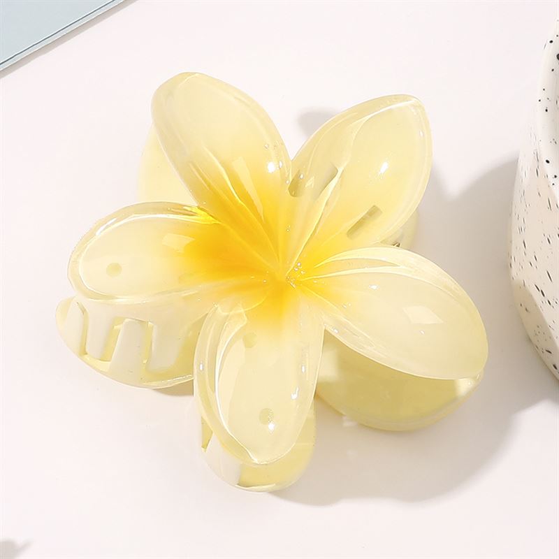 The SOHO Moana Hair Claw - Yellow, a translucent five-petaled flower accessory by SOHO, rests on a white surface beside a speckled cup.