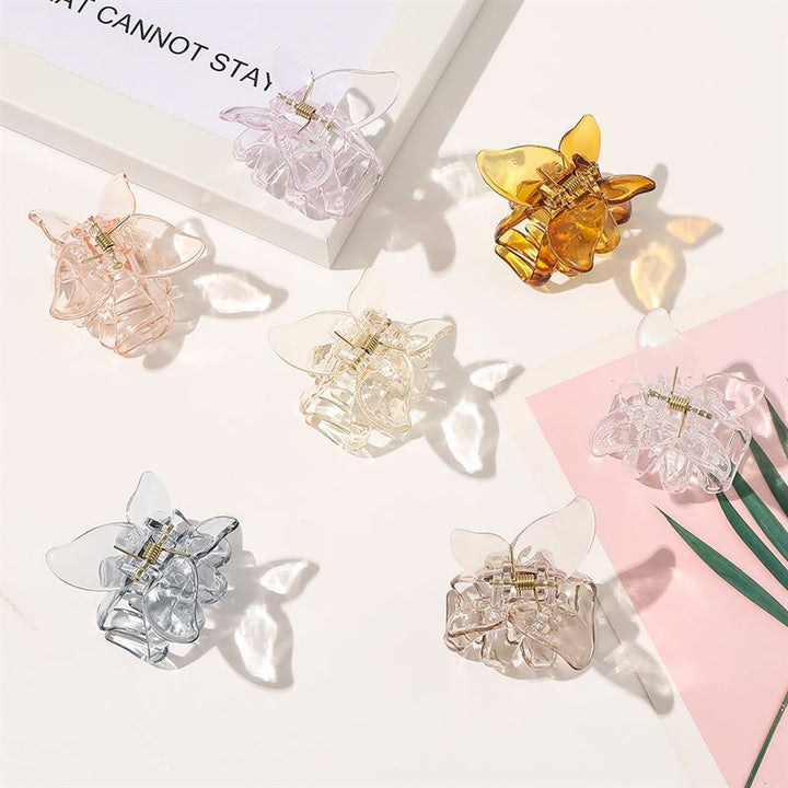 The SOHO Naja Hair Claw in Espresso by SOHO is a translucent butterfly-shaped hair accessory in a rich transparent brown shade, shown on a white surface with subtle plant leaves and papers for an elegant touch.