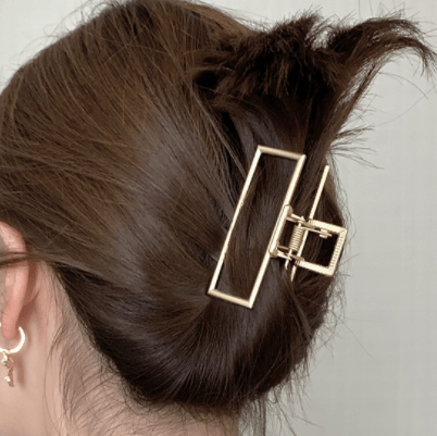 A person with brown hair in a secure messy bun held by the SOHO Niba Large Metal Hair Claw in Gold. Their ear, adorned with small earrings, is partially visible against a neutral, out-of-focus background.