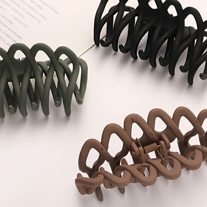 The SOHO Olive Hair Claw - Brown by SOHO, with a matte finish and visible spring, features a wavy interlocking design that provides a gentle grip for all hair types. It rests stylishly on a light surface beside an open book.
