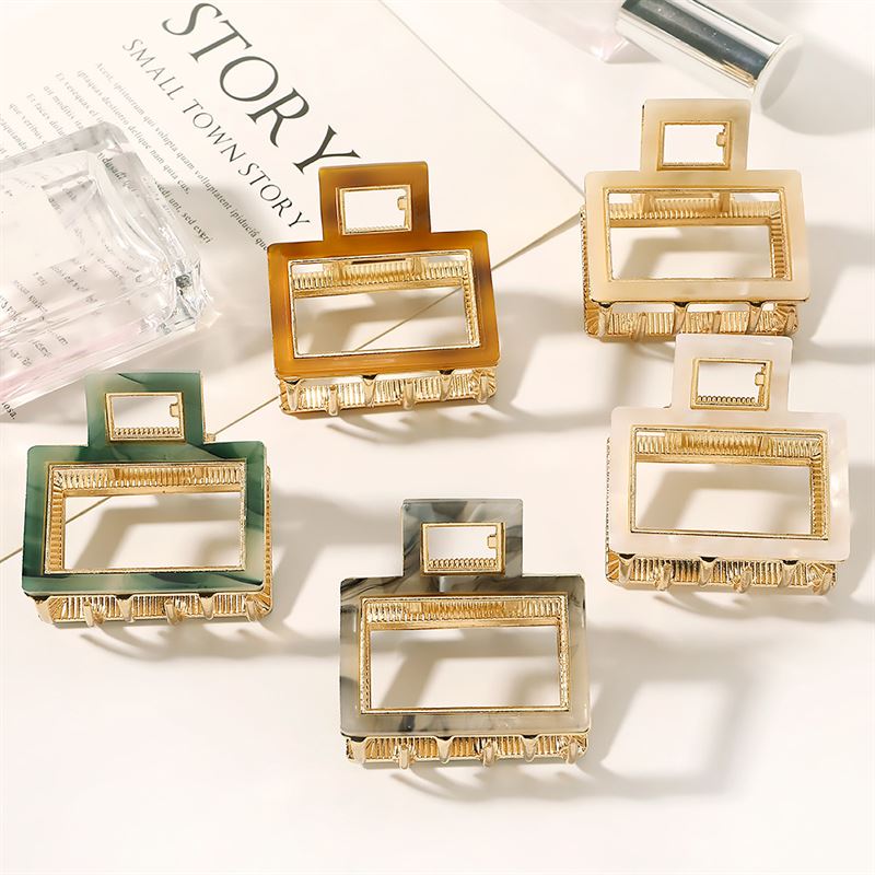 The SOHO Vida Hair Claw in Brown by SOHO is displayed alongside other geometric metal hair clips on a light surface with a decorative magazine and a clear acrylic accessory nearby.