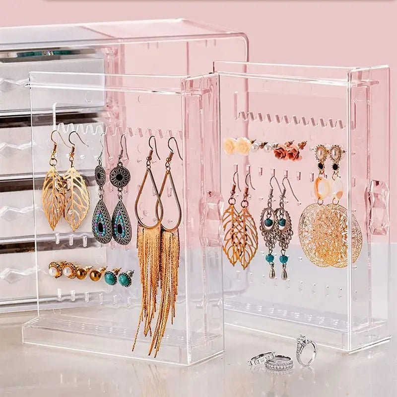 The UNIQ Acrylic Jewellery Organiser with 4 drawers & 2 earring holders (SF 1142) by UNIQ displays gold, turquoise, and feather earrings, plus a pair of rings in front, all set against a pink background.