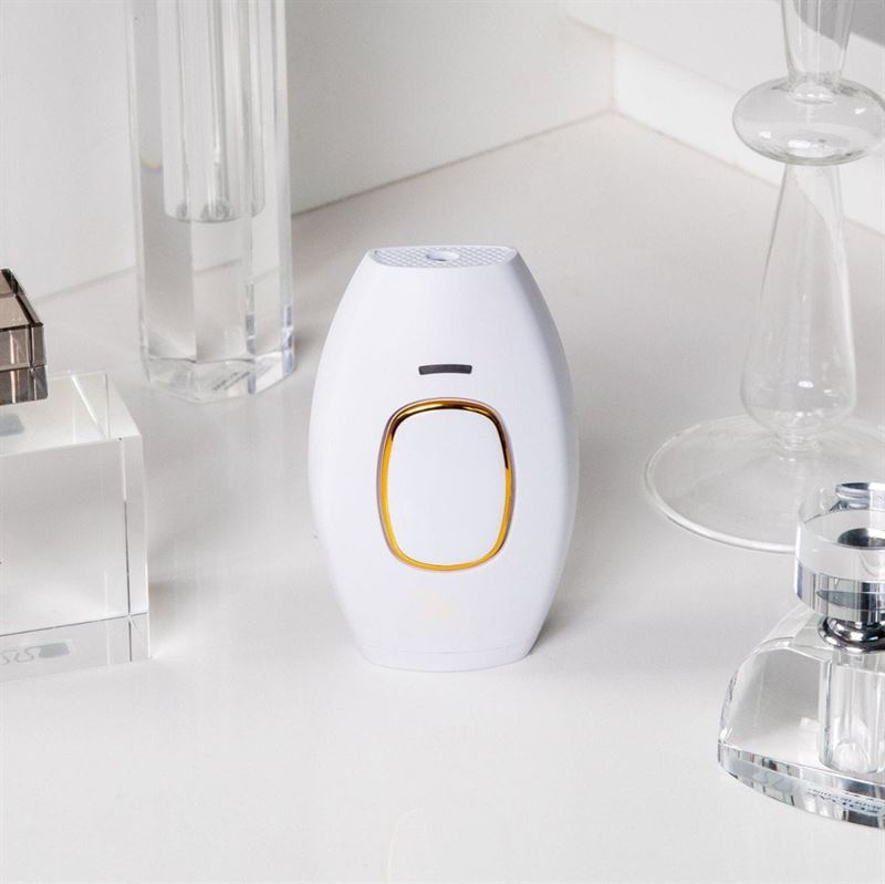 The UNIQ IPL Hair Removal Device in white, with a gold-bordered button, rests on a white countertop among glass decor—offering smooth, pain-free permanent hair reduction at home with 600,000 flashes.