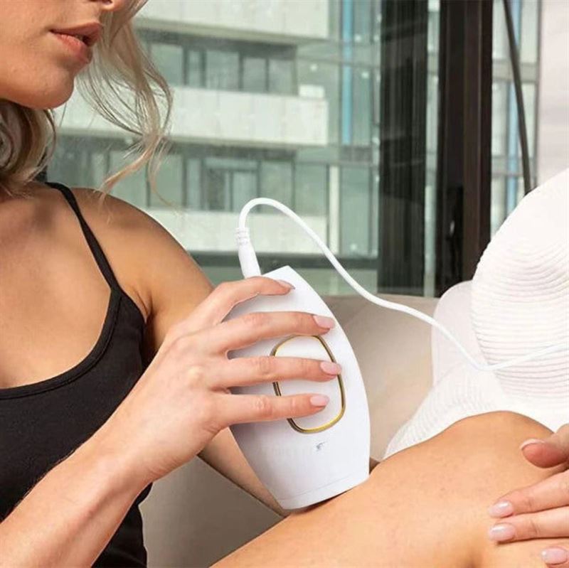 A blonde woman uses the UNIQ IPL Hair Removal Device—offering smooth, pain-free permanent hair reduction at home—on her leg while sitting indoors in a black tank top. The device is plugged in, with large windows in the background.