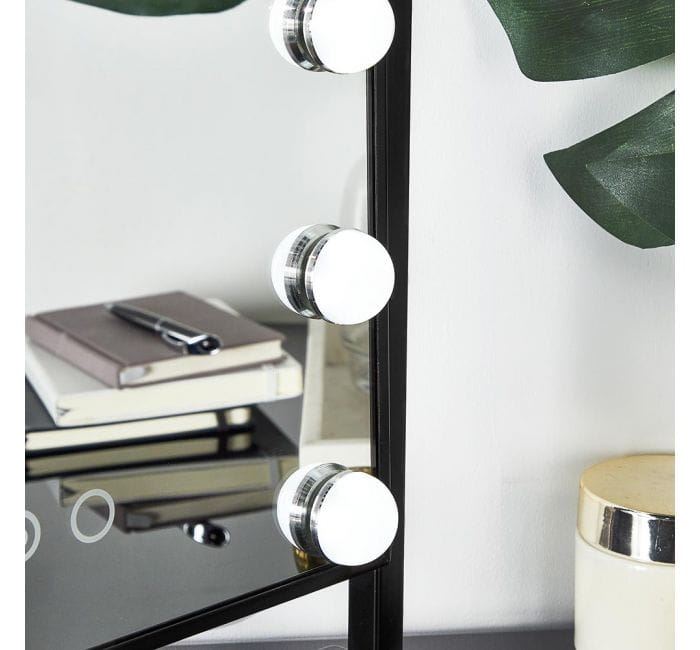 A close-up of the UNIQ New York Mega Makeup Mirror with 12 bulbs in black, by UNIQ, is shown on a dark tabletop beside stacked books, a closed pen, green leaves, and a lidded container.