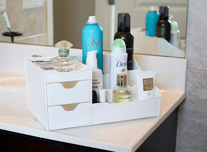 A UNIQ Organiser for makeup & skincare products with 9 compartments in white holds perfume bottles, lotion, spray cans, a tube, and other personal care items on a bathroom counter with a mirror and sink in the background.